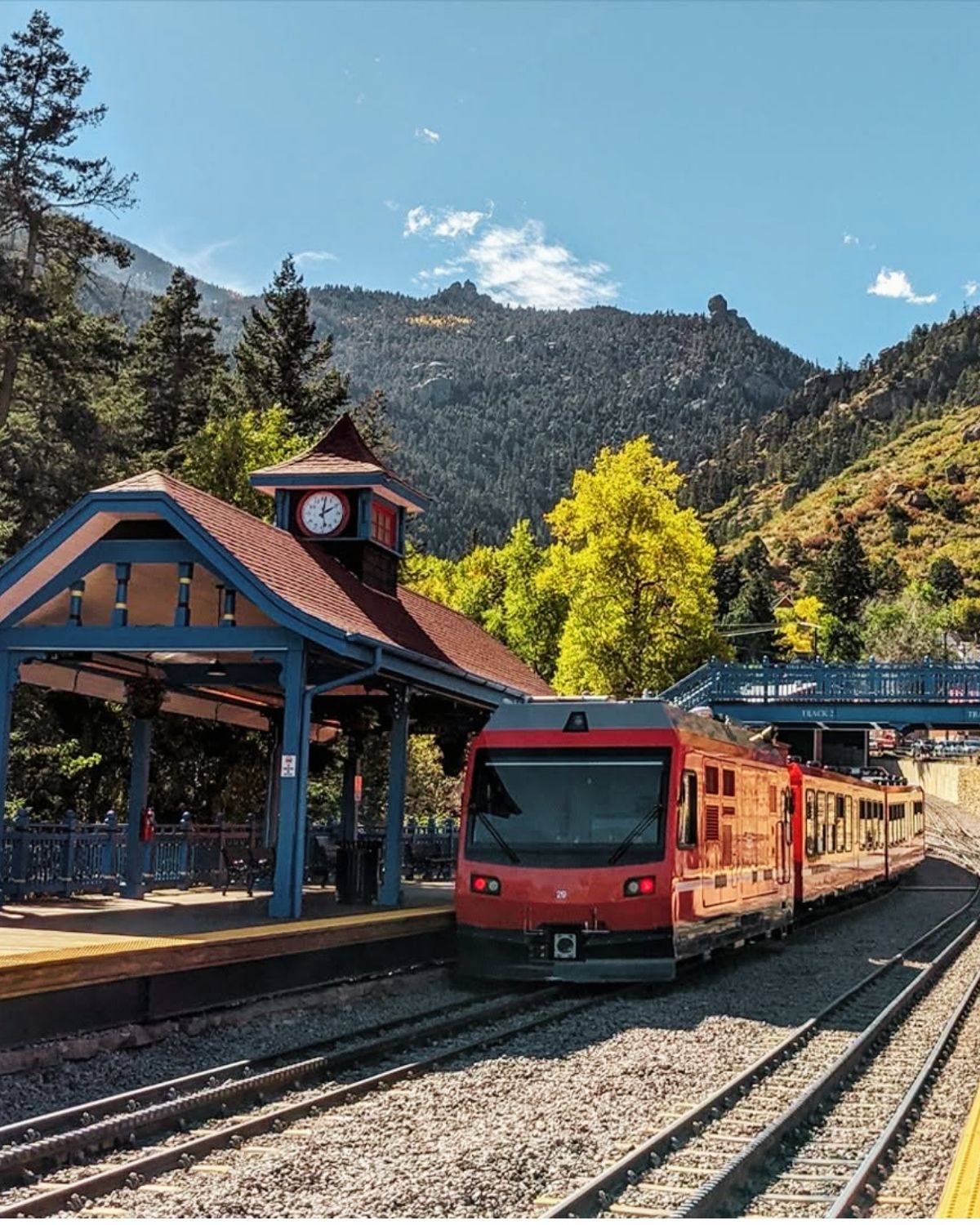 A red Pikes Peak Cog Railway train waiting at the Manitou Springs depot under its blue timber canopy, wooded slopes rising behind