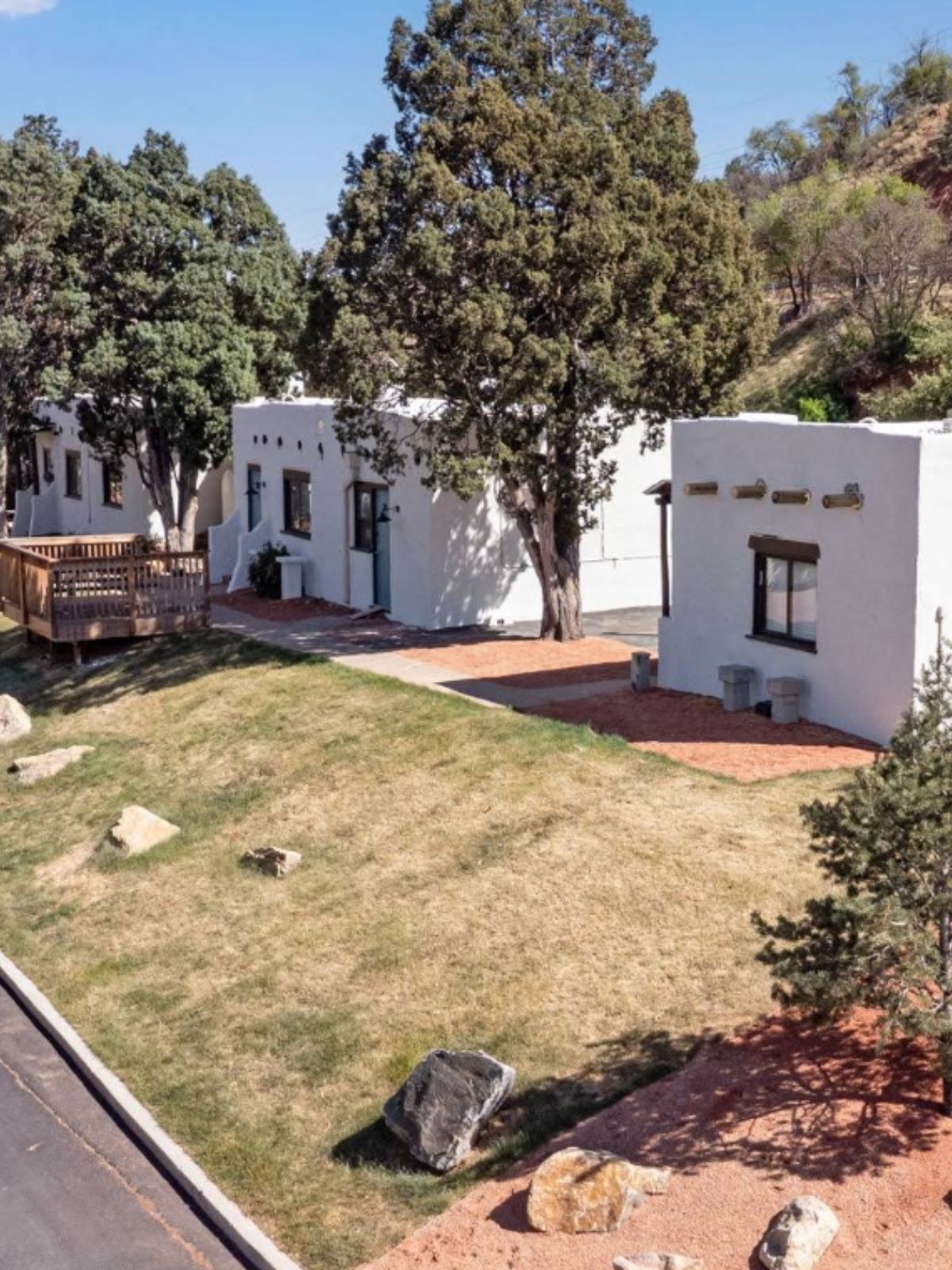 Three freestanding white adobe casitas standing apart on the lawn among juniper and pine, with red gravel paths between them and the foothills rising behind