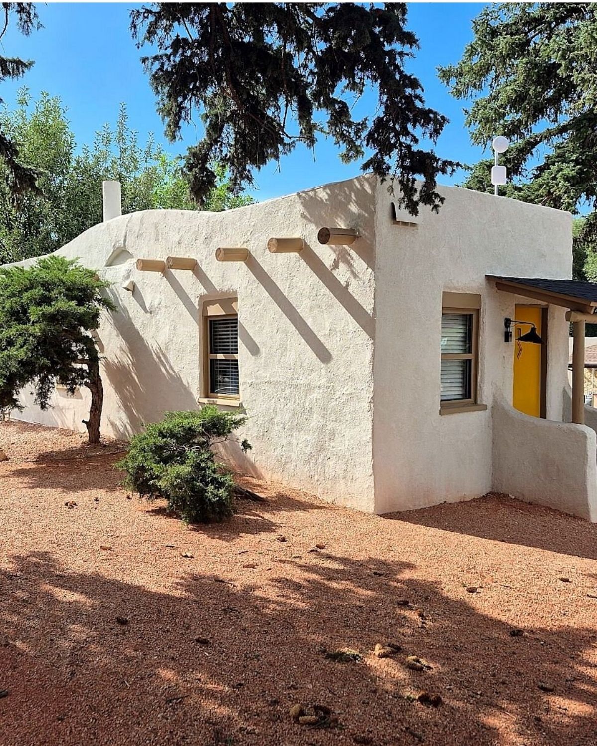A freestanding white adobe casita with peeled log vigas through the parapet and a yellow front door, junipers and red gravel around it