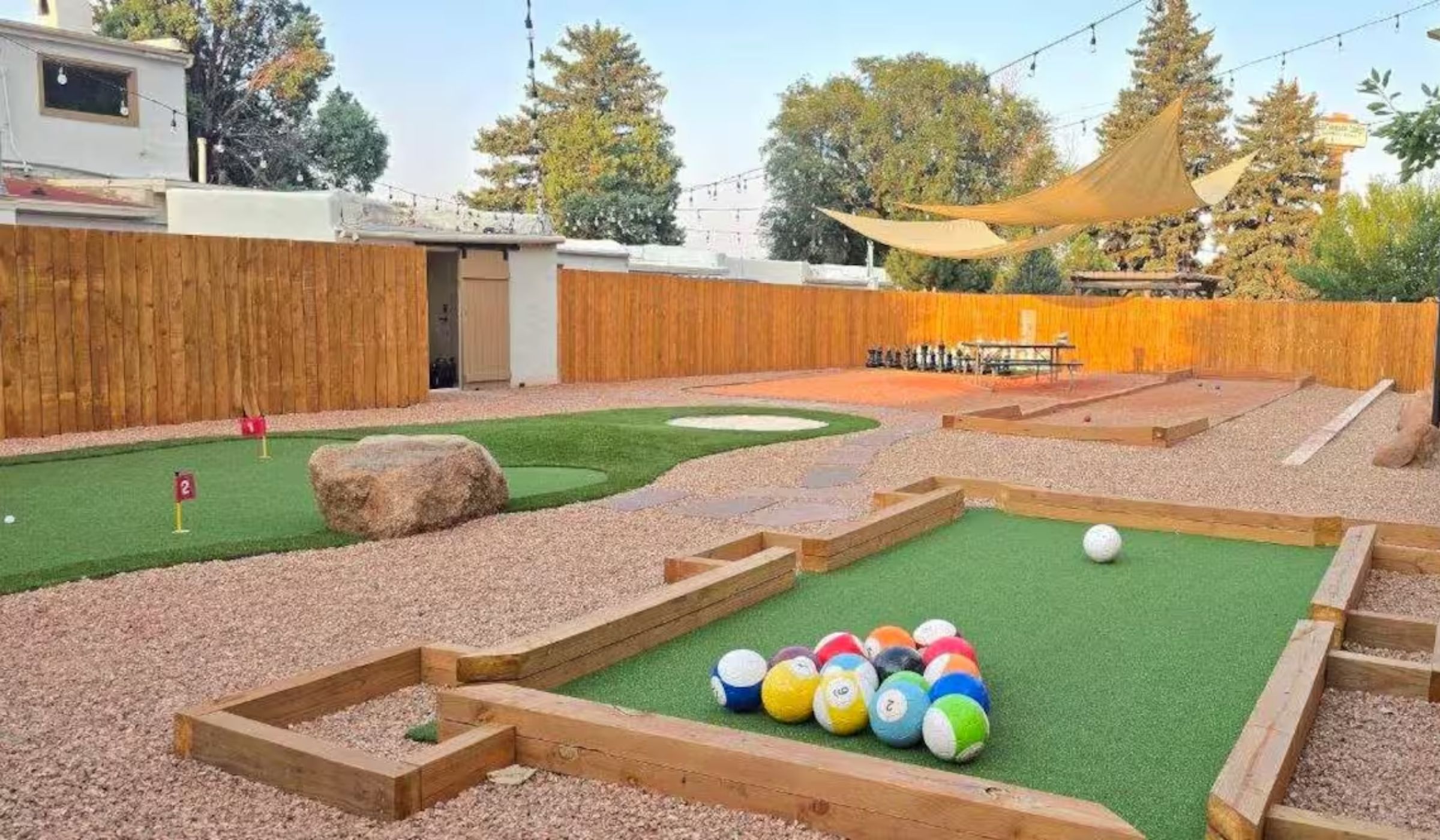 The game area at dusk: a putting green with numbered flags, a bocce court, giant chess and a ball green, under string lights and shade sails inside a cedar fence