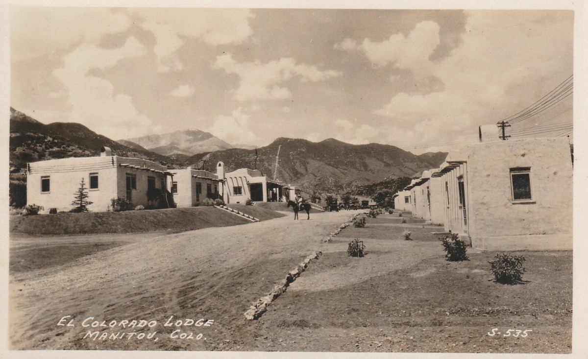 A 1920s real-photo postcard captioned El Colorado Lodge, Manitou, Colo., looking down the dirt drive between two rows of adobe casitas, with a man on horseback in the middle of the road and the mountains beyond