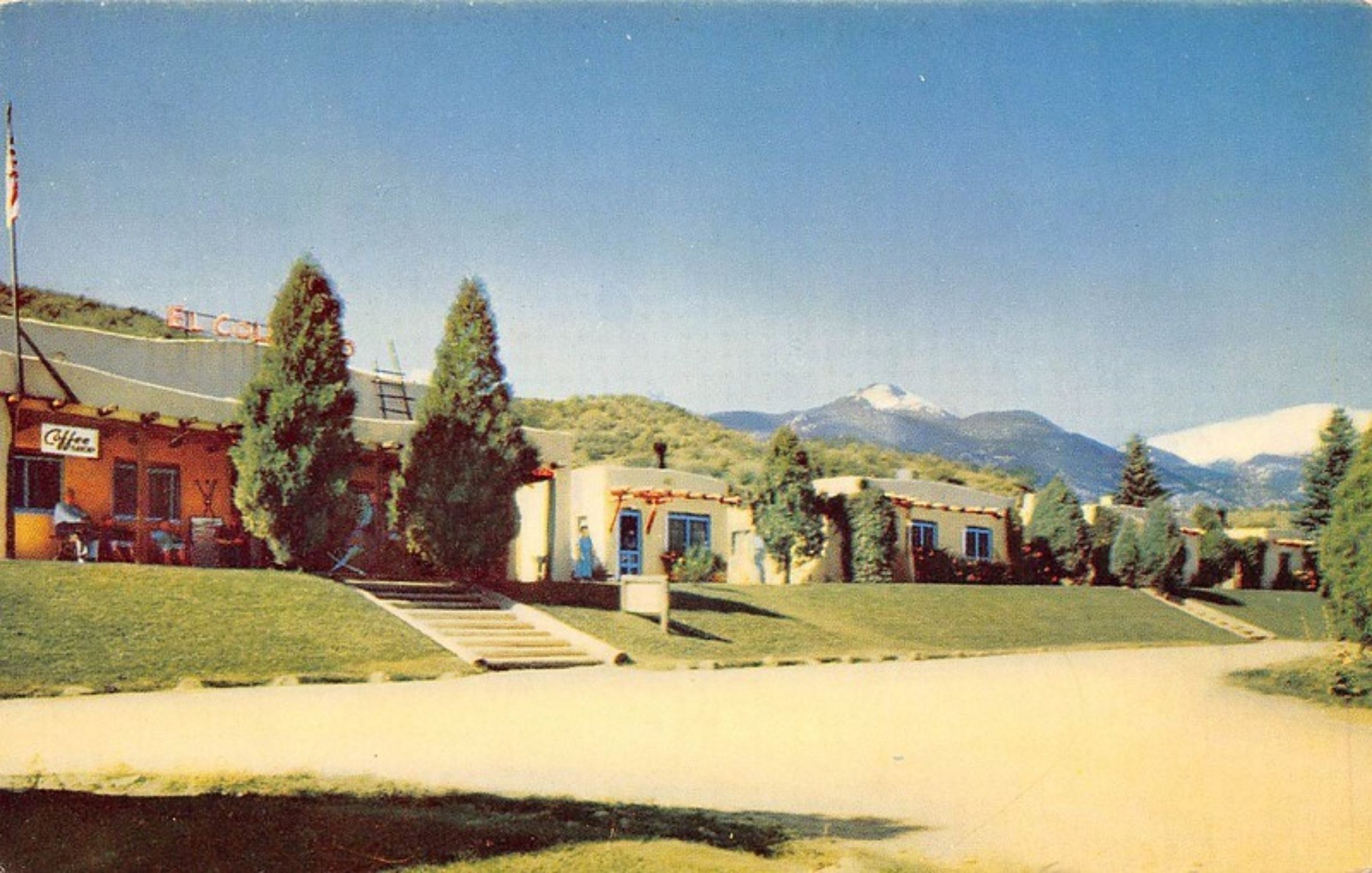 A colour postcard of El Colorado from a later era: the neon roof sign above the lodge, a Coffee Shop sign on the near building, casitas with blue doors along a clipped lawn, and the snow on Pikes Peak beyond
