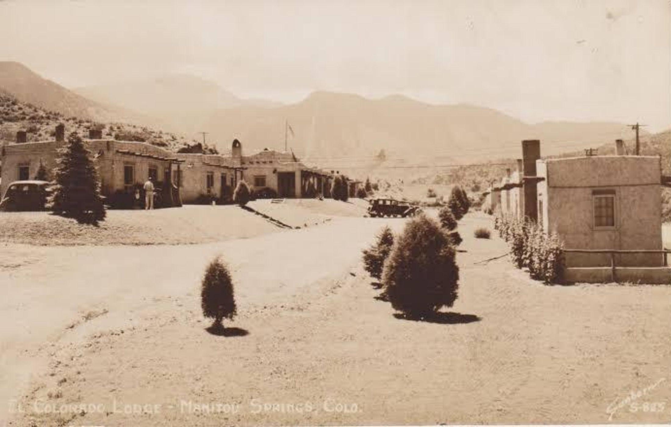 A sepia real-photo postcard captioned El Colorado Lodge, Manitou Springs, Colo., looking across the gravel drive to the low adobe cabins and the main lodge, a period automobile parked at the near cabin and the hills of Ute Pass behind