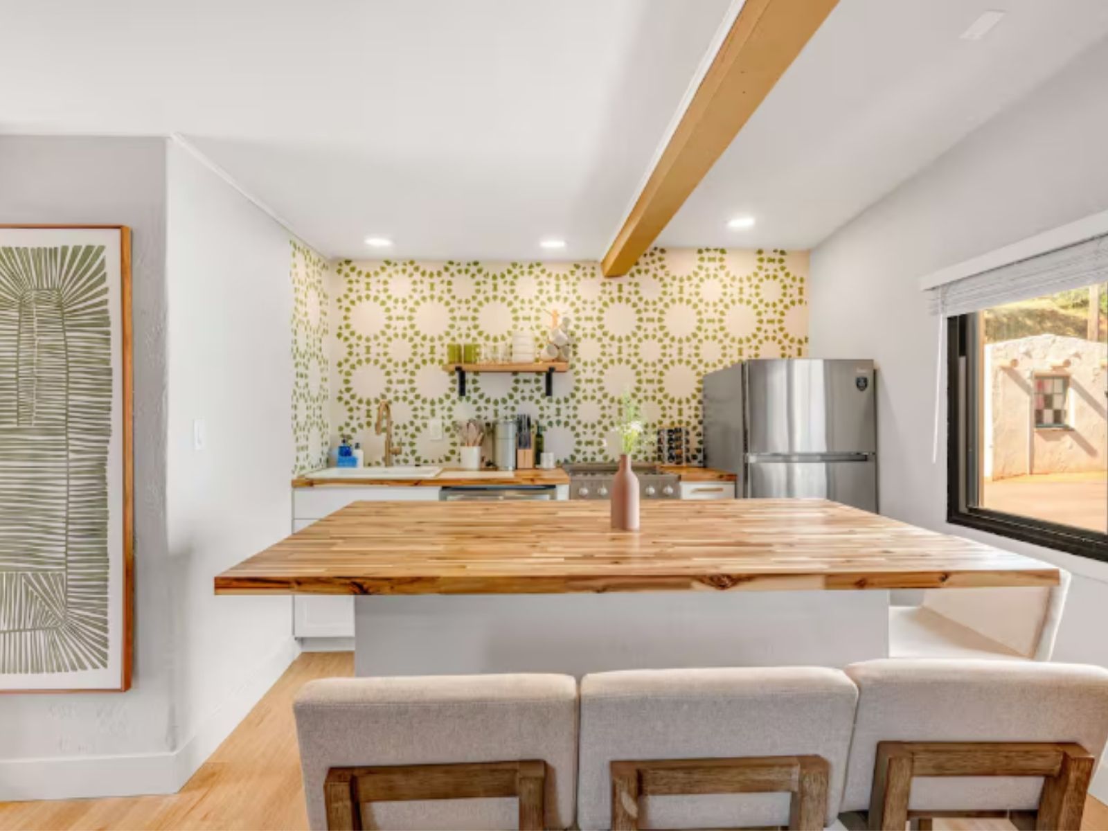 The same kitchen after restoration: white cabinetry, a butcher block island with stools, green patterned wallpaper, open shelving and the original viga left exposed overhead